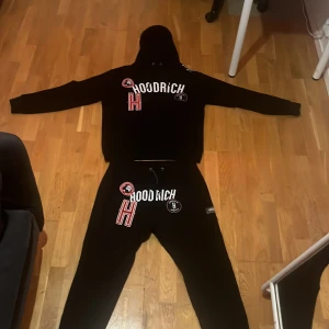 Svart hoodie från Hoodrich - Svart hoodie från Hoodrich med tryck och broderade detaljer i vitt och rött på bröstet. Stor huva med dragsko och ribbade muddar vid ärmslut och nederkant. Snygg streetwear-stil med tydlig logga framtill.