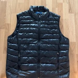 Svart dunväst från Polo Ralph Lauren - Snygg svart dunväst från Polo Ralph Lauren med hög krage och dragkedja framtill. Västen har två fickor med dragkedja och quiltad design som ger en sportig och stilren look. Perfekt att bära över en hoodie eller tröja.