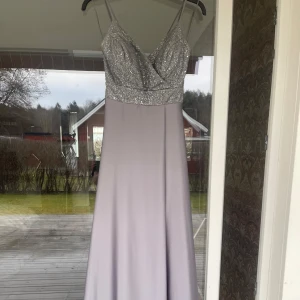 Ljuslila långklänning med glittrig överdel - Säljer en elegant ljuslila/grå långklänning med glittrig, paljettprydd överdel och smala axelband. Klänningen har v-ringning, snörning i ryggen och en slät, flowy kjol. Perfekt för bal eller festliga tillfällen. Storlek M, passar mindre!