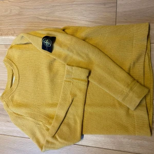 Gul stickad tröja från Stone Island - Säljer en gul stickad tröja från Stone Island med klassisk patch på ärmen. Tröjan har rund halsringning och långa ärmar. Perfekt för dig som gillar streetwear och vill ha något unikt i garderoben.