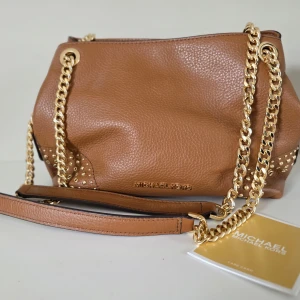 Brun axelväska från Michael Kors med guldkedja - Dags att skiljas från det som inte kommer till användning hos mig. Brun axelväska från Michael Kors i skinn med guldfärgade kedjor och detaljer. Väskan har flera fack med dragkedja och logotyp framtill. Prislapp kvar, se bild. Se även andra fina väskor i min profil
