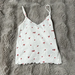 Vit ribbad linnetopp med jordgubbar från H&M - Supersöt vit ribbad linnetopp från H&M med små röda jordgubbsmönster över hela. Toppen har tunna axelband och vågig kant nertill. Perfekt för varma dagar eller som ett lager under en skjorta.