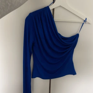  One shoulder topp - Säljer en kornblå topp med one shoulder-design och draperad framsida. Toppen har en lång ärm och en smal axelrem på ena sidan. Perfekt för dig som vill sticka ut med en trendig och färgstark look!