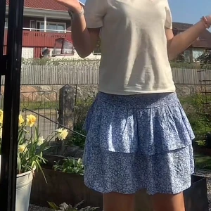 Blå blommig volangkjol - Supersöt blå kjol med vitt blommönster och volanger. Kjolen har en smockad midja som gör den bekväm och sitter snyggt. Perfekt för varma dagar och ger en lätt och luftig känsla.