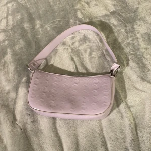 Ljuslila axelväska med präglat mönster - Säljer en ljuslila axelväska med präglat mönster och silvriga detaljer. Väskan har en enkel design med dragkedja upptill och ett kort axelband. Perfekt för att bära det viktigaste och ge din outfit en söt touch.
