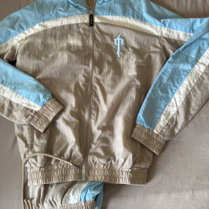 Trapstar Tracksuit  - Säljer en beige och ljusblå Tracksuit från Trapstar med dragkedja och resår vid ärmslut och midja. Jackan har en broderad Trapstar-logga på bröstet och sportig design med kontrastpaneler. Perfekt för dig som gillar streetwear. Storlek S M L XL XXL är tyvärr sålda men de finns en sista bit av storlek XS passa på och köp först till kvarn!