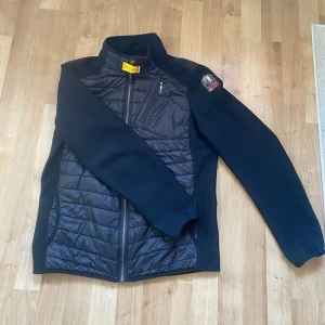 Svart hybridjacka från Parajumpers - Säljer en svart hybridjacka från Parajumpers med quiltad front och rygg i blankt material samt stickade ärmar och sidor. Jackan har dragkedja framtill, hög krage och en bröstficka med dragkedja. Klassisk PJS-logga på ärmen.