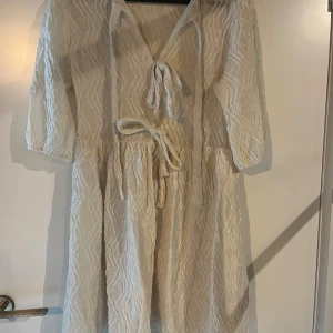 Vit klänning med knytband - Säljer en vit klänning med strukturmönster och v-ringning. Klänningen har trekvartsärm och knytband både i midjan och vid halsen. Perfekt för sommarens tillfällen och har en luftig känsla. 