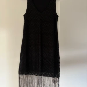Svart klänning med fransar  - Säljer en svart ärmlös klänning från Amisu i storlek 36. Klänningen har ett snyggt sicksackmönster och långa fransar nertill som ger en cool retrovibe. Perfekt för dig som vill sticka ut med en unik look!