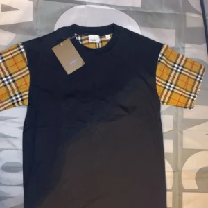 Svart t-shirt med rutiga ärmar - Säljer en svart t-shirt från Burberry med klassiska beige-rutiga ärmar och matchande detalj vid halsen. T-shirten har en normal passform och är kortärmad. Perfekt för dig som gillar stilrena plagg med en twist.