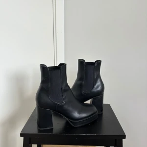 Svarta chelsea boots med klack - Säljer ett par svarta chelsea boots i skinnimitation med hög klack och plattform. Skorna har elastiska paneler på sidorna och rund tå. Perfekta för att lyfta din outfit.