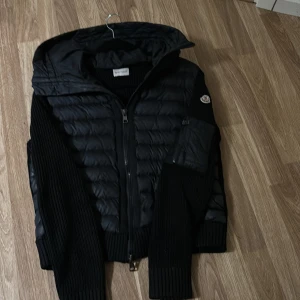 Svart pufferjacka från Moncler - Säljer en svart pufferjacka från Moncler med stickade ärmar och huva. Jackan har dragkedja framtill och Moncler-logga på ena ärmen. Perfekt för kyliga dagar och riktigt snygg streetstil. Jackan är äkta.