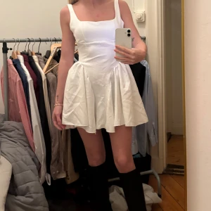 Vit kortklänning med rosett i ryggen - Supersöt vit klänning med breda axelband och markerad midja. Kjolen är utställd och har mycket vidd, och baktill finns en stor rosett som detalj. Perfekt för sommarens alla tillfällen!