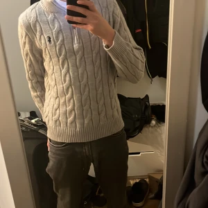 Ljusgrå kabelstickad tröja från Polo Ralph Lauren - Säljer en ljusgrå kabelstickad tröja från Polo Ralph Lauren. Tröjan har halv dragkedja vid halsen, ribbade muddar och klassisk broderad logga på bröstet. Perfekt för dig som gillar stilrena och tidlösa plagg. Priset sitter inte i sten och kan diskuteras 