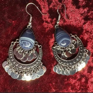 Bohemiska örhängen med blå sten, silverdetaljer och vintageinspirerad design - Vackra örhängen i bohemisk stil med droppformad blå sten och dekorativa silverfärgade detaljer. Den mönstrade stenen ramas in av en halvmåneformad metallstruktur med små hängande berlocker som ger ett fint och elegant rörelsemönster när du bär dem. Lätta och bekväma – perfekta för dig som gillar boho, orientalisk stil eller vill ha ett smycke som sticker ut.  Längd: ca 6,5 cm Material: Silverfärgad metall, blå sten Skick: Mycket bra begagnat skick Ingen prutning – bundlerabatt erbjuds!  