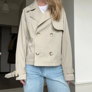 Beige kort trenchcoat med bälte vid ärmslut - Snygg beige trenchcoat med dubbelknäppning och bred krage. Jackan är croppad och har bältesdetaljer vid ärmsluten. Perfekt för en stilren look till våren eller hösten. (Lånade bilder) obs! Går tyvärr inte ner i pris då intresset är stort.