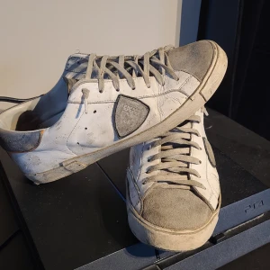 Vita och grå sneakers med grå detaljer - Säljer ett par vita phillipe model med grå detaljer och snörning. Skorna har en rund tå och platt sula, samt en tydlig logotyp på sidan. Perfekta för en avslappnad och sportig stil. Allt og förekommer