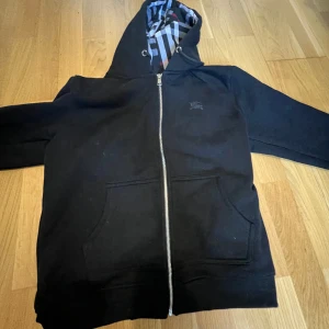 Burberry Kofta - Säljer en svart hoodie från Burberry med dragkedja och klassiskt rutigt foder i huvan. Tröjan har två stora fickor framtill och en broderad Burberry-logga på bröstet. Perfekt för en stilren och bekväm look. Storlek L men passar M