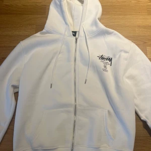Vit Stüssy Zip Hoodie  - Säljer en vit zip hoodie från Stüssy. Tröjan har svart Stüssy-logga tryckt på bröstet och stor logga med text på ryggen.  Aldrig använd, endast testad. Nypris ca 2000kr.