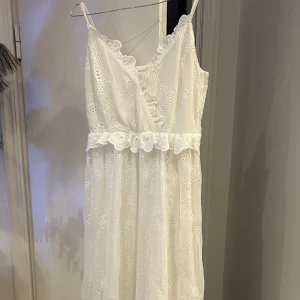 Vit spetsklänning med broderade blommor - Romantisk vit klänning med smala axelband och vacker spetsdetalj. Klänningen har broderade blommor, volang i midjan och omlottliknande v-ringning. Perfekt för sommarens tillfällen eller student.