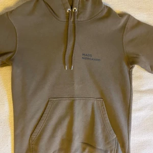 Olivgrön hoodie från Mads Nørgaard - Jag säljer en olivgrön hoodie i storlek S från Mads Nørgaard med diskret logga på bröstet. Tröjan har huva med dragsko och en stor magficka framtill. Liten fläck på magfickan.