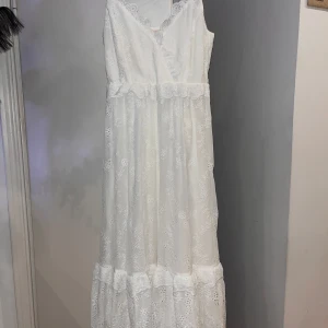VitStudent klänning med spetsdetaljer  - Romantisk vit långklänning med v-ringning och vackra spetsdetaljer. Klänningen har ärmlös design och markerad midja med volangkant. Perfekt för sommarens speciella tillfällen. 
