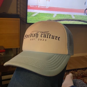 Ljusblå och vit keps med texten 'Swedish culture EST. 2023' - Snygg keps i ljusblått och vitt med svart tryck framtill där det står 'Swedish culture EST. 2023'. Kepsen har mesh-bakstycke och justerbar snapback. Perfekt accessoar för en avslappnad stil.