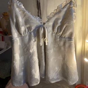 Vit blommig linnetopp med volang - Söt vit linnetopp med blommigt mönster och volangkant. Toppen har smala axelband och knytning framtill. Perfekt för sommardagar och ger en romantisk känsla till outfiten. Sitter verkligen super fin på en, använd ksk 3-4 gånger bara🩵