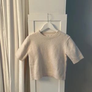 Beige stickad tröja från Kappahl - Mysig beige stickad tröja från Kappahl med korta ärmar och ribbade muddar vid hals, ärmslut och nederkant. Perfekt för lager-på-lager och enkel att matcha med olika outfits.