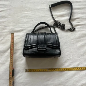 Svart axelväska i krokopräglat mönster - Snygg svart axelväska med krokopräglat mönster och kedjerem. Väskan har ett dekorativt spänne framtill och ett litet handtag upptill. Perfekt för att bära det viktigaste och ger en cool touch till outfiten.