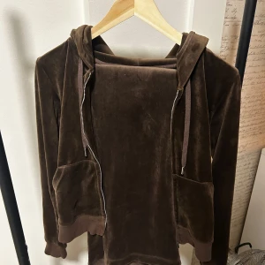Brun velour hoodie och byxa från Juicy Couture  - Mjuk och skön brun hoodie och byxa i velour från Juicy Couture. Tröjan har dragkedja, huva med snören och två fickor framtill. Byxan har snören med silver detaljer längst ut. Samt på baksidan på rumpan finns det två fickor med knappar som man stänger med.  Perfekt för en avslappnad och trendig look.
