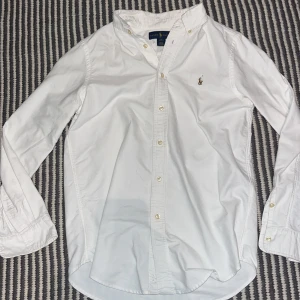 Vit skjorta från Ralph Lauren - Klassisk vit skjorta från Ralph Lauren med broderad logga på bröstet. Skjortan har lång ärm, knappar framtill och en normal passform. 