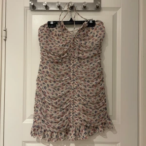 Blommig kortklänning med volang från Zara - Supersöt kortklänning från Zara med blommigt mönster i flera färger. Klänningen har smala axelband som knyts i nacken, rynkade detaljer framtill och volang längst ner. Perfekt för sommarens alla tillfällen!