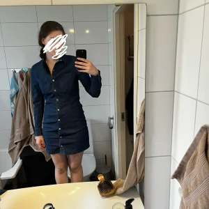 Mörkblå jeansklänning med knappar - Säljer en mörkblå jeansklänning med långa ärmar och knappstängning hela vägen framtill. Klänningen har en klassisk skjortkrage och två bröstfickor. Snygg och enkel modell som passar perfekt till vardags. 🌺
