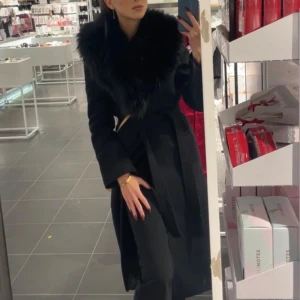 Svart kappa med fuskpäls - Säljer en svart lång kappa med stor krage i fuskpäls och knytskärp i midjan. Jackan har lång ärm och en elegant siluett, perfekt för dig som vill ha en stilren look.