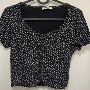 Svart blommig croppad topp - Svart croppad topp med små vita blommor och v-ringning. Toppen har korta ärmar och knappar framtill. Perfekt för en trendig och avslappnad stil.