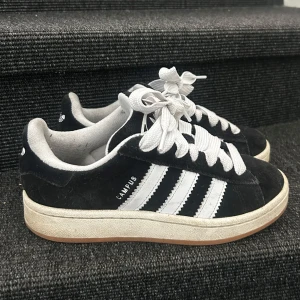 Svarta Adidas Campus sneakers - Klassiska svarta Adidas Campus sneakers med vita ränder och vit sula. Skon har en låg profil, rund tå och är tillverkad i mocka med tydliga vita snören. Perfekt för dig som gillar en stilren och tidlös look. Använda 5 gånger