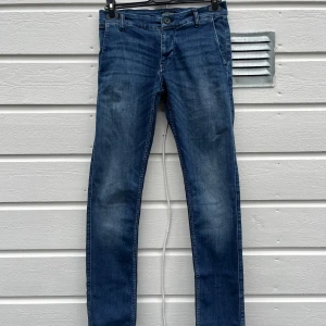 Blå denimbyxor från Lindbergh - Snygga blå denim chinos från Lindbergh i mjuk, följsam stretch denim ”SUPERFLEX” med eleganta, smakfulla detaljer - perfekt för en stilren look. Otroligt bra skick, endast använda två gånger och tvättade en gång.