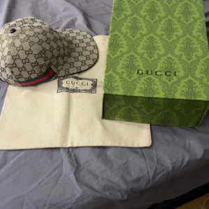 Beige keps från Gucci med monogram - Säljer en beige keps från Gucci med klassiskt GG-monogram och grönt/rött band på sidan. Kepsen har böjd skärm och är tillverkad i canvasmaterial. Kommer med originalpåse och låda. Säljes billigt vid snabbare afärr 