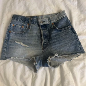 Levi's 501 jeansshorts  - Klassiska blå jeansshorts från Levi's 501 med mid waist och råa, fransiga kanter. Shortsen har slitningar framtill och femficksdesign. Perfekta för sommaren och ger en avslappnad vintagekänsla. Storlek W25. Nypris 800kr. 