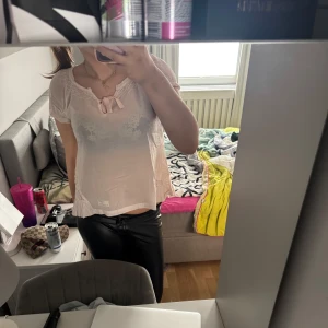 Ljusrosa blus med spets och rosett - Söt ljusrosa blus med spetsdetaljer och en liten rosett framtill. Blusen är kortärmad och har en feminin, romantisk stil. Perfekt att matcha med både jeans och kjol.