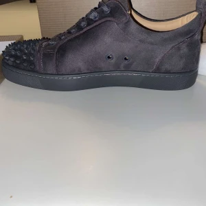 Grå Christian Louboutin skor - Säljer ett par svarta sneakers i mocka från Louboutin med coola svarta nitar på tån. Skorna har snörning och en platt sula, perfekt för dig som vill sticka ut med din stil. Storlek 43 pris ej hugget i sten! 