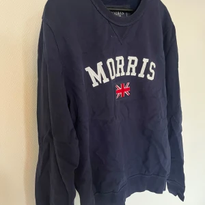 Mörkblå sweatshirt från Morris - Mörkblå sweatshirt från Morris med broderad logga och brittisk flagga på bröstet. Klassisk rund halsringning och långa ärmar. Perfekt för en stilren och avslappnad look.