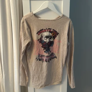 Beige långärmad tröja med tryck från Zadig & Voltaire - Säljer en beige långärmad tröja från Zadig & Voltaire med stort tryck av döskalle, blommor och text på framsidan. Tröjan har råa kanter vid halsringningen och en avslappnad passform. Perfekt för en cool och avslappnad stil.