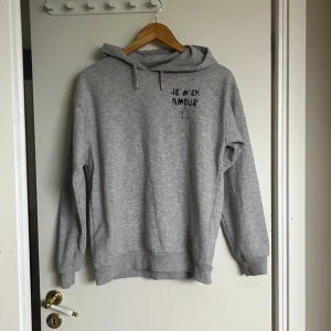 Grå hoodie från Gina Tricot med tryck - Säljer en ljusgrå hoodie från Gina Tricot med texten 'JE M'EN AMOUR' och en stjärna tryckt både fram och bak. Tröjan har huva och långa ärmar, perfekt för en avslappnad stil.