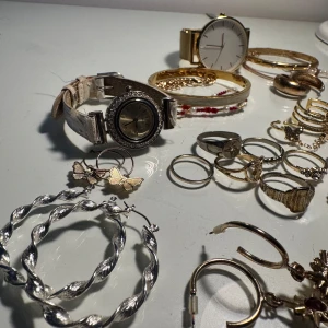 Mix av smycken och klockor i silver och guld - Samling med flera smycken: armband, ringar, halsband och örhängen i både silver- och guldfärg. Inkluderar två armbandsur, pärlhalsband, örhängen med pärlor, stjärnhänge och glittriga detaljer. Perfekt för dig som gillar att mixa olika stilar och material.