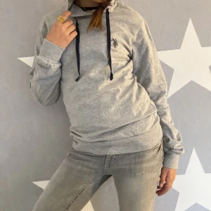 Grå hoodie från U.S. Polo Assn. - Säljer en ljusgrå hoodie från U.S. Polo Assn. med mörkblå dragsnören och tryckt logga på bröstet. Tröjan har huva och långa ärmar, perfekt för en avslappnad stil. Materialet är mjukt och skönt bomullsmix.