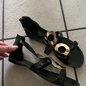 Svarta sandaler med gulddetaljer från Paulo Boccelli  - Snygga svarta sandaler från Carlo Bacelli med stora runda guldfärgade metalldetaljer framtill. Skorna har dragkedja bak och remmar över foten. Perfekta för dig som vill sticka ut med en elegant touch.