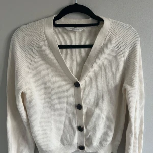 Vit ribbad kofta med knappar - Säljer en  vit kofta från H&M med ribbad struktur och stora svarta knappar framtill. Koftan har lång ärm och v-ringad halsringning, perfekt att slänga över en topp eller klänning. Den har en liten gul fläck på axeln, inte så märkbar. Använt flera gånger. Pris kan diskuteras!🩷