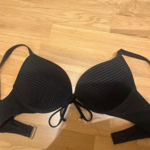 Svart ribbad bikiniöverdel från H&M - Svart bikiniöverdel från H&M med ribbad struktur och knyt mellan kuporna. Har guldfärgade detaljer på banden och justerbart spänne i ryggen. Klassisk form med vadderade kupor och smala axelband.